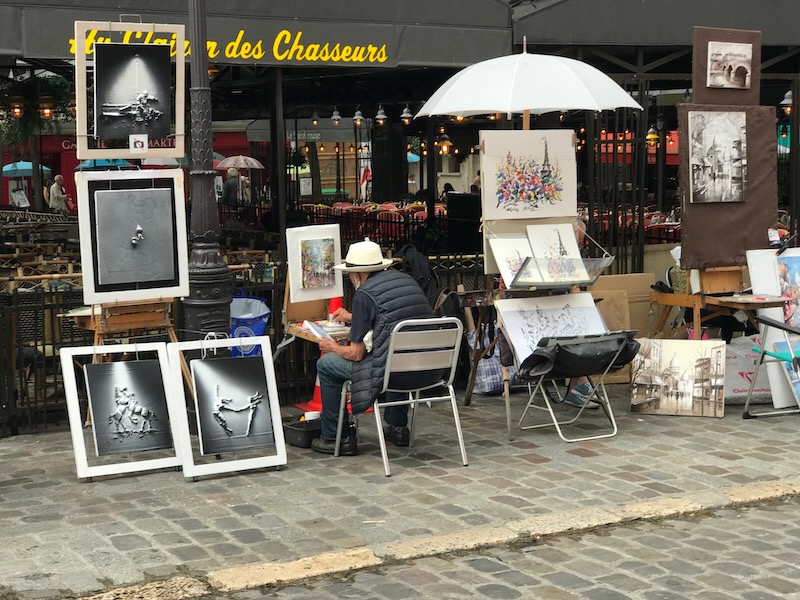 man sitting on chair on the side of the road  in Montmartre painting canvas and surrounded by paintings 