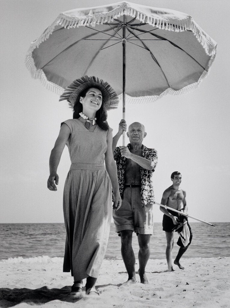 A black-and-white photograph of Pablo Picasso on a beach, wearing an unbuttoned printed shirt and shorts and holding a sun umbrella for Françoise Gilot, who walks in front of him in a long dress and beach hat. A shirtless young man walks behind them.